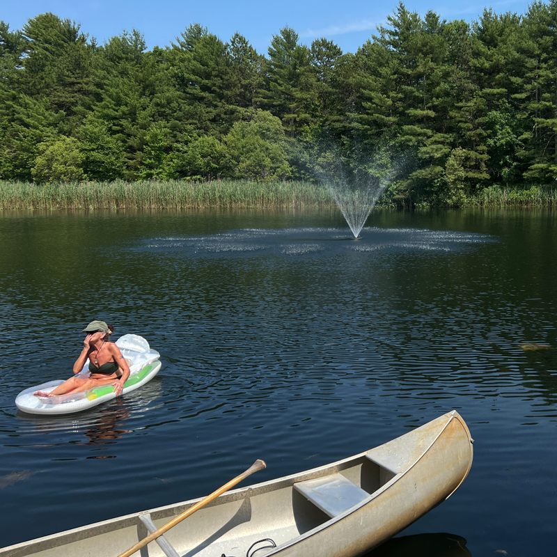 Person relaxing on a float in a lake surrounded by lush trees with a fountain in the center, next to a canoe.