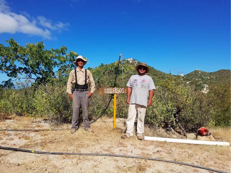 Two men wearing hats standing in front of a Los Pisos sign in a rural landscape with trees and hills under a clear blue sky.