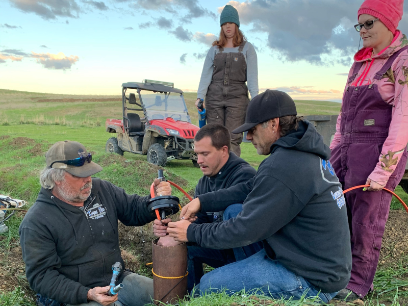 Group of people working on a water well installation in a grassy field with an ATV in the background under a cloudy sky.